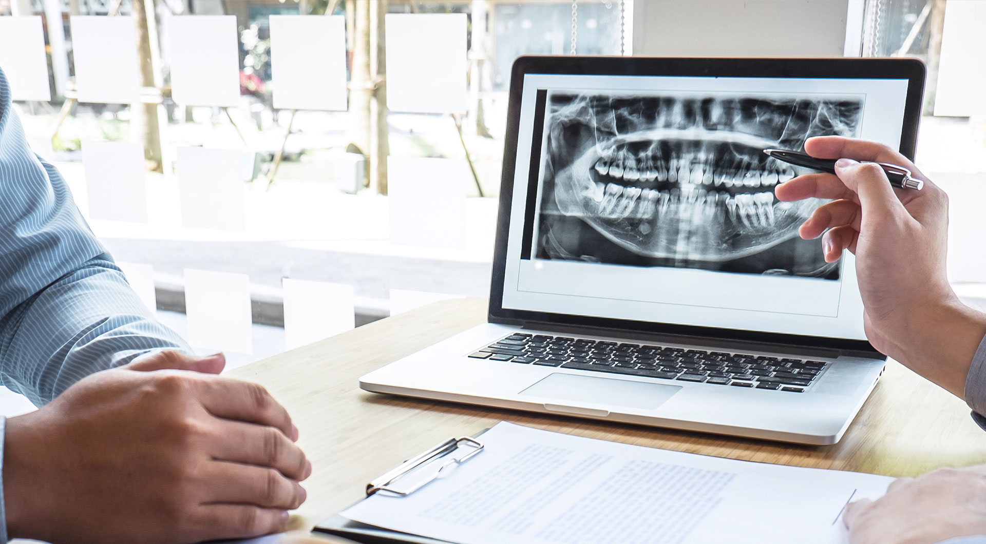 A split-screen image showing two different scenes  on the left, a person working at a laptop with a presentation screen displaying an X-ray of a human jaw  on the right, a close-up view of someone s hands pointing to the same X-ray displayed on the laptop.