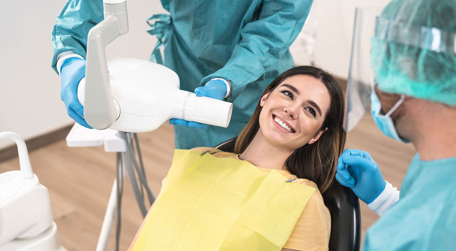 A woman receiving dental treatment from a professional using a dental chair, with a hygienist standing behind her, both wearing protective gear.